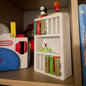 May include: A white bookshelf with three shelves, each holding small books and figurines. The top shelf has a panda figurine and a red dinosaur figurine. The middle shelf has a white puppy figurine. The bottom shelf has four small books with titles "Dancing Dinos", "Poky Little Puppy", "The Little Red Hen", and "The Very Busy Spider".