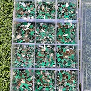 Puede incluir: Un contenedor de almacenamiento de plástico transparente con doce compartimentos individuales llenos de pequeñas piezas de espejo reflectantes en varias formas geométricas, incluyendo gotas, diamantes y círculos, cada una con un reverso verde.