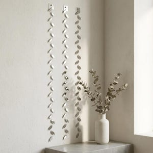 Puede incluir: Tres guirnaldas verticales de pared compuestas por una serie de pequeños elementos reflectantes en forma de hoja de color plateado unidos a un hilo fino. Cada tira está anclada en la parte superior por una placa cuadrada plateada. Un jarrón blanco contiene ramas de eucalipto.