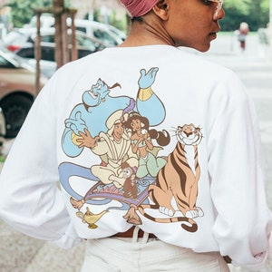 May include: White long-sleeve t-shirt with a graphic print of Aladdin, Jasmine, Genie, Abu, and Rajah on a magic carpet. The characters are in a cartoon style and are colored in shades of blue, green, brown, and yellow.