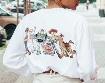May include: White sweatshirt featuring a colorful graphic of characters from the animated movie Toy Story. The design includes Woody, Buzz Lightyear, Jessie, and other characters. The sweatshirt has long sleeves and a crew neck.