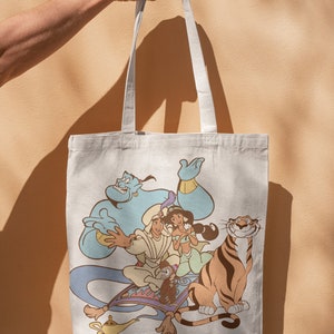 May include: A white canvas tote bag with a colorful print of Aladdin, Jasmine, Abu, and Rajah from the Disney movie Aladdin. The characters are on a magic carpet flying through the air.