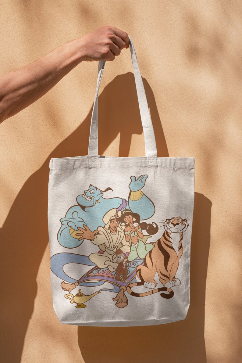 May include: A white canvas tote bag with a colorful print of Aladdin, Jasmine, Abu, and Rajah from the Disney movie Aladdin. The characters are on a magic carpet flying through the air.