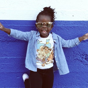 May include: A young person wearing a white t-shirt with a cartoon image of Aladdin and Jasmine on a magic carpet. The t-shirt is layered under a blue denim jacket. The person is wearing sunglasses and smiling.