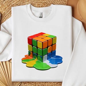 May include: White sweatshirt with a vibrant, melting Rubik's Cube graphic. The cube's squares are red, orange, yellow, green, and blue, with the colours dripping down the front. The sweatshirt has a crew neck and long sleeves.