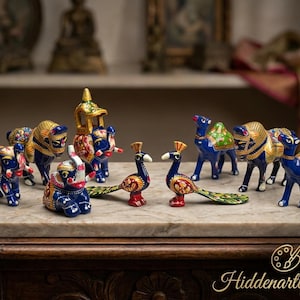 May include: A collection of hand-painted animal figurines including elephants, camels, and peacocks. The figures feature vibrant blue, red, and green colors with intricate gold detailing. The text Hiddenartbyram is visible in the bottom right corner.