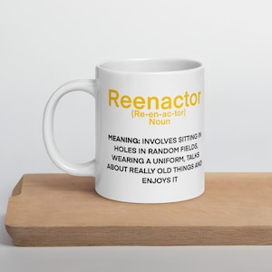 May include: White ceramic mug featuring the word "Reenactor" in yellow, with a definition of the hobby. The definition is humorous. The mug is placed on a wooden surface.