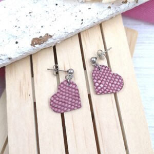 May include: A pair of pink heart-shaped earrings with a textured surface. The earrings are made of leather and have silver-toned metal hooks.