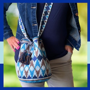 May include: A crochet bucket bag featuring a geometric diamond pattern in shades of navy blue, light blue, and brown. The bag includes a matching woven shoulder strap and a drawstring closure finished with two decorative tassels at the ends.