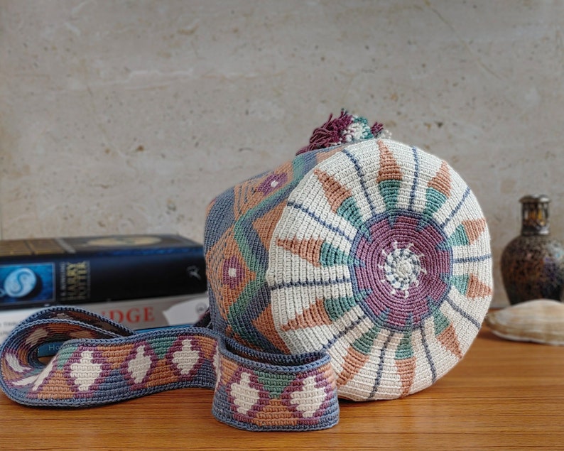 May include: A cylindrical crochet bag featuring a circular base with a sunburst pattern in cream, mauve, teal, and terracotta. The body and long strap display intricate geometric diamond motifs in matching colors, finished with a small tassel on top.