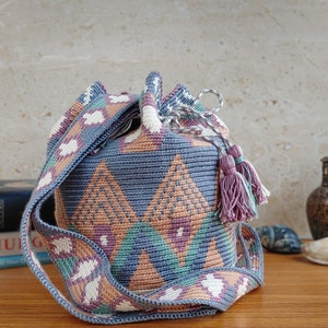 May include: A cylindrical crochet bucket bag featuring a geometric pattern in shades of dusty blue, peach, teal, and cream. The bag includes a wide, matching patterned shoulder strap and a drawstring closure finished with two decorative yarn tassels.