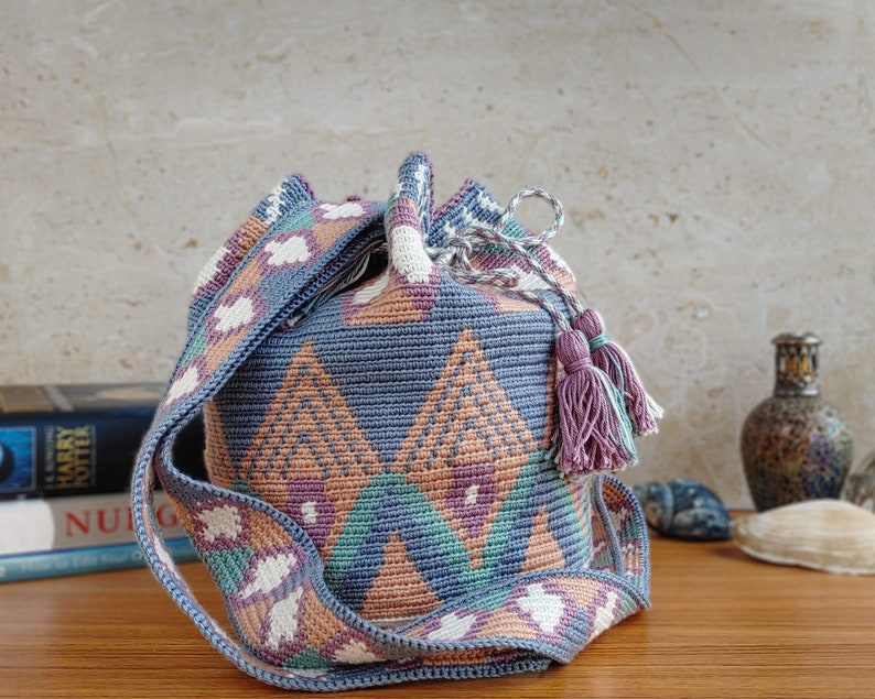 May include: A cylindrical crochet bucket bag featuring a geometric pattern in shades of dusty blue, peach, teal, and cream. The bag includes a wide, matching patterned shoulder strap and a drawstring closure finished with two decorative yarn tassels.