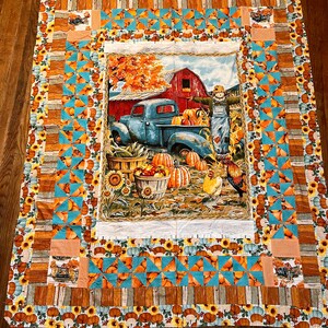 Puede incluir: Una colcha de retazos con temática otoñal. El panel central presenta un camión vintage, un granero rojo, un espantapájaros y calabazas. El borde está hecho de tela azul y naranja con un diseño de calabaza y girasol.