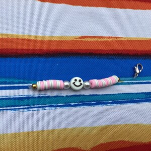 Puede incluir: Un brazalete de cuentas blancas y rosadas con un cierre dorado y un dije de carita sonriente blanca.