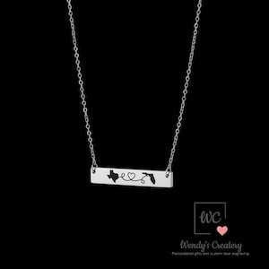 May include: A silver-colored necklace with a rectangular pendant. The pendant features the state outlines of Texas and Florida, connected by a heart-shaped design. The necklace is suspended by a delicate chain.