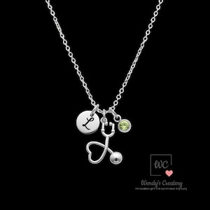 May include: A silver necklace with a stethoscope charm, a round charm with the letter "L", and a small green gemstone. The stethoscope has a heart-shaped chest piece. The necklace hangs against a black background.