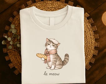 May include: A white t-shirt featuring a watercolor illustration of a gray tabby cat wearing a beret and scarf, holding a baguette. The text "le meow" is written below the cat.