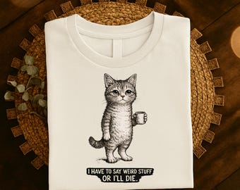 Op de afbeelding: Crèmekleurig T-shirt met een zwart-wit illustratie van een kat die een koffiekopje vasthoudt. De kat staat rechtop. Onder de afbeelding staat de tekst: "I HAVE TO SAY WEIRD STUFF OR I'LL DIE."