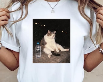 Puede incluir: Camiseta blanca con un meme de un gato sentado con una botella de agua. El gato es gris y blanco, con una expresión aburrida. El fondo es oscuro con algunas luces.