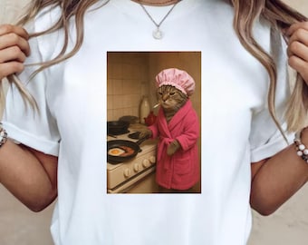 May include: White t-shirt with a framed image of a cat in a pink dressing gown and cap, smoking a cigarette while cooking eggs. The cat is in front of a stove with a frying pan. The image has a vintage, humorous style.