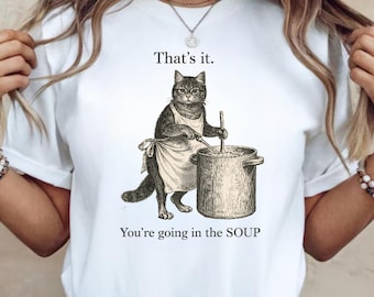 Könnte beinhalten: Weißes T-Shirt mit einer Vintage-Illustration einer Katze, die eine Schürze trägt und in einem Topf rührt. Der Text "That's it. You're going in the SOUP" ist über und unter dem Katzenbild gedruckt.
