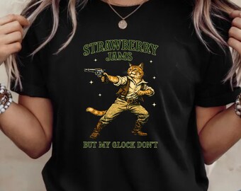 Peut inclure: T-shirt noir avec un chat de dessin animé vêtu d'une tenue de cow-boy, tenant un pistolet. Le texte "STRAWBERRY JAMS" est au-dessus du chat, et "BUT MY GLOCK DON'T" est en dessous. Le chat est orange et beige.