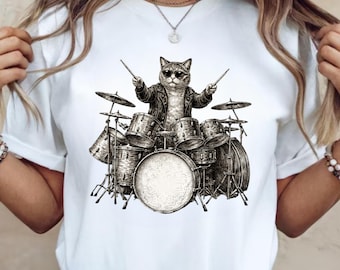 May include: White t-shirt featuring a black and white illustration of a cat wearing sunglasses and a leather jacket, playing a drum set. The cat is holding drumsticks, with a full drum kit visible.