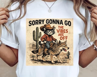 Peut inclure: T-shirt blanc avec un motif d'un chat portant un chapeau de cowboy chevauchant un loup, avec le texte "SORRY GONNA GO THE VIBES ARE OFF". Le motif comprend des cactus et un paysage désertique.
