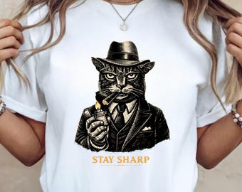 Könnte beinhalten: Weißes T-Shirt mit einer detaillierten Illustration einer Katze in Anzug und Hut, die eine Zigarre raucht und ein Feuerzeug hält. Der Text "STAY SHARP" ist darunter in Gold gedruckt.
