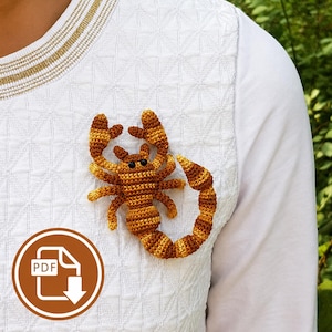 May include: A crocheted scorpion brooch in shades of golden yellow and brown, featuring two large pincers, eight legs, and a curved tail with a stinger. The scorpion has two small black bead eyes and is attached to a white textured shirt.
