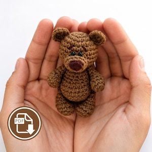 Könnte beinhalten: Ein kleiner, brauner, gehäkelter Teddybär mit einer dunkelbraunen Nase und schwarzen Perlenaugen. Der Bär hat eine strukturierte Oberfläche aus Garn, abgerundete Ohren und eine kompakte, sitzende Haltung mit sichtbaren Maschen.