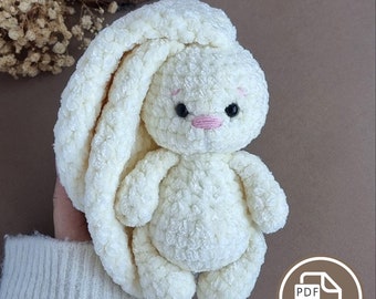 Pode incluir: Coelho de crochê branco com orelhas compridas, nariz rosa e olhos pretos. O coelho está sentado em uma mão que está usando um suéter branco.