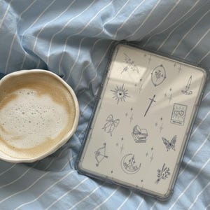 May include: A white ceramic mug filled with a cappuccino sits next to a tablet with a clear case. The tablet case has a white background with various small, blue illustrations. The items rest on a blue and white striped bedsheet.