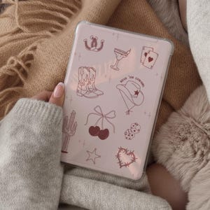 以下が含まれることがあります： ピンクのカウボーイをテーマにしたデザインの透明なタブレットケース。デザインには、蹄鉄、カウボーイブーツ、カクテル、トランプ、カウボーイハット、さくらんぼ、サイコロ、サボテン、星、ハートが含まれています。「Read 'em Cowgirl」というテキストも含まれています。