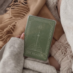 May include: A green book cover featuring a sword and sun design, with the words "THE READER". The book is held in a hand, resting on a blanket. The cover has an ornate border.