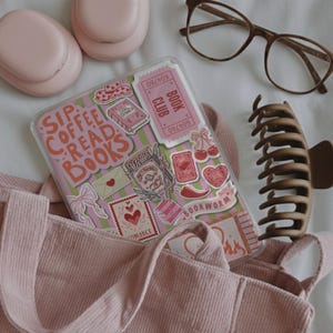 May include: A pink corduroy tote bag with a clear phone case decorated with stickers that read "Sip Coffee Read Books". Other items include pink earbuds, brown glasses, and a brown hair clip. The theme is centered around reading and books.