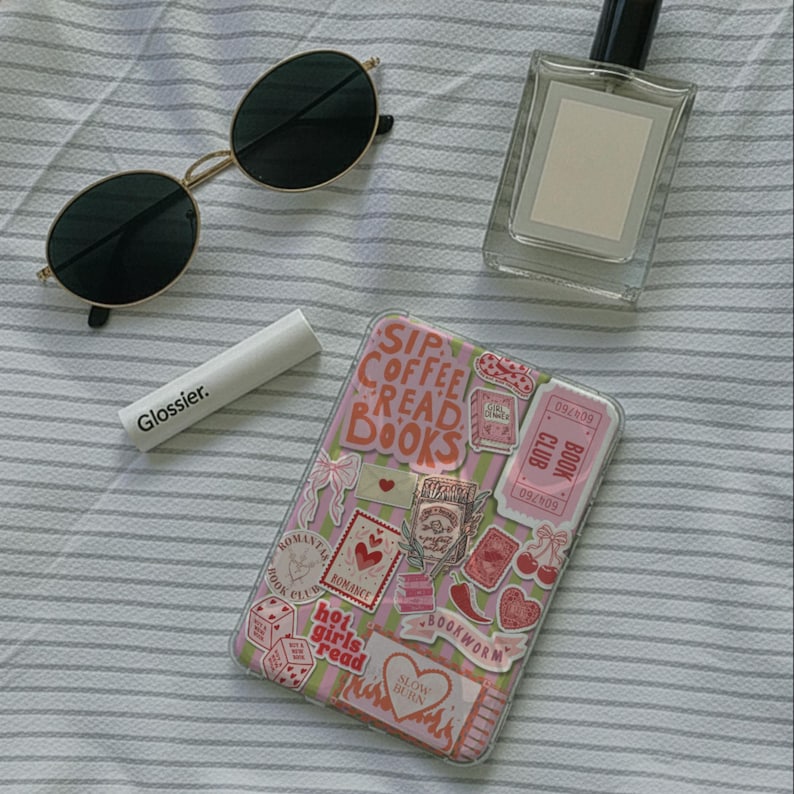 May include: An overhead shot of a collection of items on a striped surface. Includes a pair of gold-rimmed sunglasses, a tube of Glossier lip balm, a perfume bottle, and a phone case decorated with stickers that read "Sip Coffee Read Books."