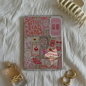 May include: A clear tablet case decorated with stickers featuring the words "Sip Coffee Read Books", hearts, and book-related imagery. A gold perfume bottle, gold hoop earrings, and a cream-colored hair clip are also present.