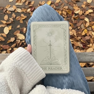 May include: A rectangular, off-white case with a celestial design featuring a sword, sun, and open book. The text "THE READER" is at the bottom. The case is held on a person's lap, with blue jeans and a white sweater visible.