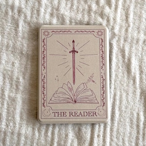 Puede incluir: Una carta de tarot con fondo crema e ilustración burdeos. La ilustración presenta una espada sobre un libro abierto, con el texto "THE READER" en la parte inferior. La tarjeta está enmarcada con detalles florales y solares.