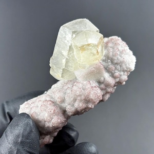 Op de afbeelding: Een groot, helder, geel calcietkristal met een complexe, gefacetteerde structuur zit bovenop een bed van wit en roze, gekristalliseerd mineraal.