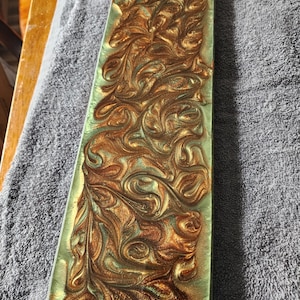 May include: A rectangular, decorative resin tray with a swirling pattern of gold and teal colors. The tray has a glossy finish and a slightly raised, textured design. The edges are smooth and rounded, creating a modern aesthetic.