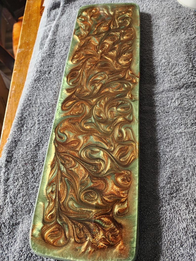 May include: A rectangular, decorative resin tray with a swirling pattern of gold and teal colors. The tray has a glossy finish and a slightly raised, textured design. The edges are smooth and rounded, creating a modern aesthetic.