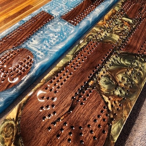May include: Two handmade cribbage boards. One board features a blue resin base with wooden playing pieces. The other board has a gold and green resin base with wooden playing pieces. Both boards have metal pegs and drilled holes for gameplay.