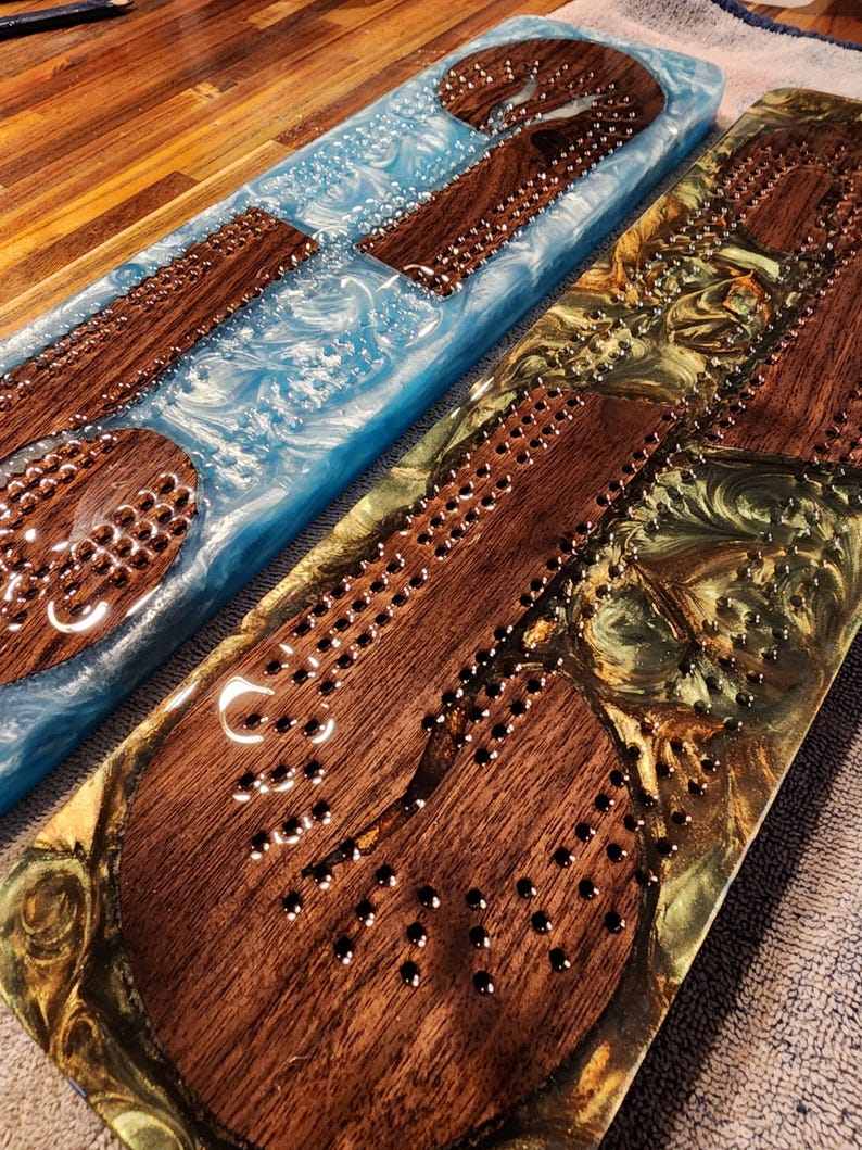 May include: Two handmade cribbage boards. One board features a blue resin base with wooden playing pieces. The other board has a gold and green resin base with wooden playing pieces. Both boards have metal pegs and drilled holes for gameplay.