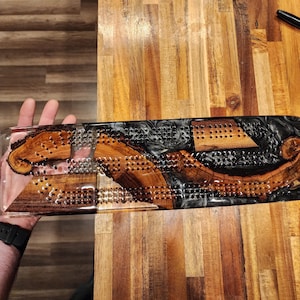 May include: A handcrafted cribbage board made of wood and resin. The board features a clear resin base with inlaid wood pieces and a black resin section. The wood has a natural grain pattern, and the board has numerous holes for pegs.
