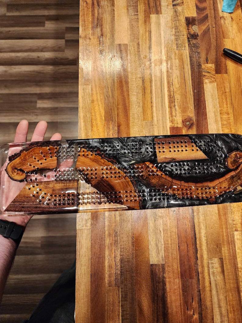May include: A handcrafted cribbage board made of wood and resin. The board features a clear resin base with inlaid wood pieces and a black resin section. The wood has a natural grain pattern, and the board has numerous holes for pegs.