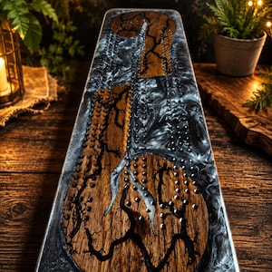 May include: A handcrafted cribbage board with a unique design. The board features a wooden surface with intricate black patterns and embedded metal pegs. The edges are encased in a swirling black and white resin, creating a striking contrast.