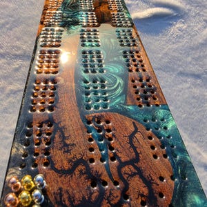 May include: A rectangular cribbage board made of dark brown wood with natural grain patterns and a glossy teal resin inlay. The board features multiple rows of drilled holes for scoring and includes a set of small metallic pegs in gold and silver.