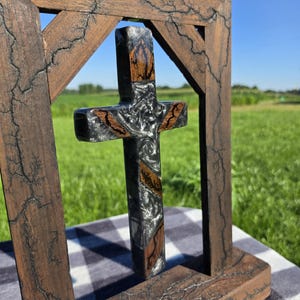 May include: A wooden cross suspended within a rectangular wooden frame. The frame features dark, lightning-like fractal burn patterns. The cross is constructed from a combination of natural brown wood segments and a swirling, metallic gray resin.